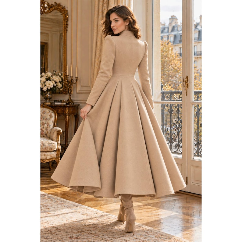May include: A long, beige, floor-length coat with a fitted bodice, structured shoulders, and a full, pleated skirt that flares out from the waist. The coat features long sleeves and a high neckline, providing a structured silhouette.