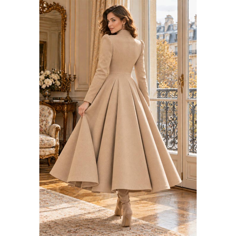 May include: A long, beige, floor-length coat with a fitted bodice, structured shoulders, and a full, pleated skirt that flares out from the waist. The coat features long sleeves and a high neckline, providing a structured silhouette.