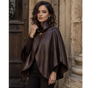 May include: A dark brown leather cape featuring a high collar and a single gold-toned metal toggle closure at the neckline. The garment has a smooth, polished finish and a wide, flowing silhouette that drapes over the shoulders and upper torso.