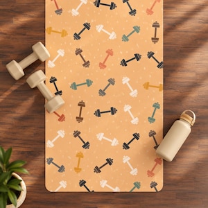Puede incluir: Una esterilla de ejercicio rectangular con fondo naranja claro que presenta un patrón repetitivo de mancuernas en varios colores, incluyendo negro, blanco, verde azulado y óxido. Dos mancuernas beige y una botella de agua beige están sobre la esterilla.