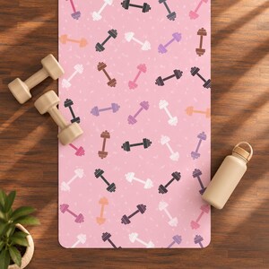 Puede incluir: Una esterilla de yoga rectangular con fondo rosa claro y un patrón repetitivo de mancuernas en varios colores, incluyendo blanco, negro, marrón, morado y naranja, acompañada de dos mancuernas beige y una botella de agua beige.