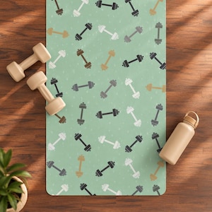 Puede incluir: Una esterilla de yoga rectangular con fondo verde claro y un patrón repetitivo de mancuernas en blanco, gris oscuro y beige. Dos mancuernas beige están colocadas en el lado izquierdo de la esterilla y una botella de agua beige a la derecha.