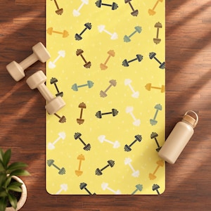 Op de afbeelding: Een rechthoekige yogamat met een gele achtergrond en een herhalend patroon van halters in diverse kleuren, waaronder wit, bruin, donkergrijs en gedempt groen. Twee beige halters en een beige waterfles liggen op de mat.