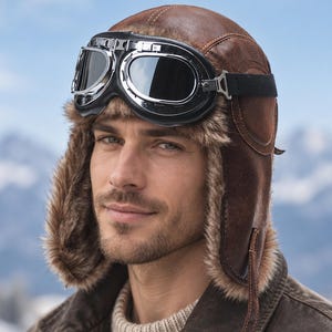 May include: A brown leather aviator hat with a soft faux fur lining and ear flaps, paired with vintage-style chrome-framed goggles featuring dark lenses. The hat has visible stitching details and a black elastic strap securing the goggles in place.