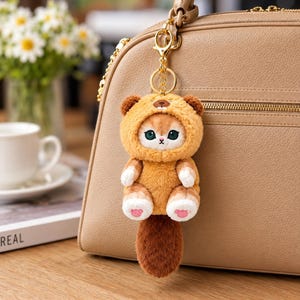 May include: A plush cat keychain featuring a soft, tan-colored hood with small ears and a tiny embroidered face. The cat has white paws with pink paw-pad details and a long, fluffy dark brown tail. It is attached to a gold-toned metal lobster clasp.