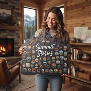 Könnte beinhalten: Ein dunkel gefärbtes, bergförmiges Holzdisplay mit dem weißen Schriftzug "Summit Stories" in der Mitte. Das Board ist mit zahlreichen kleinen, bunten, geformten Aufnähern bedeckt, die verschiedene Berglandschaften und Szenen darstellen.