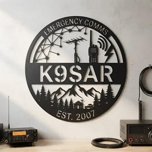 May include: A circular black metal wall sign featuring the text EMERGENCY COMMS at the top, K9SAR in the centre, and EST. 2007 at the bottom. The design includes silhouettes of a radio antenna, a handheld radio, mountains, trees, and a vehicle.