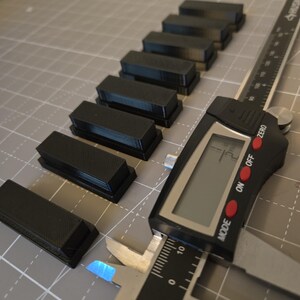 May include: Seven small, rectangular black plastic blocks arranged in a diagonal line. A digital caliper is positioned next to the blocks, displaying a measurement of 1.2 inches on its screen, with the metal jaws open to measure one of the blocks.