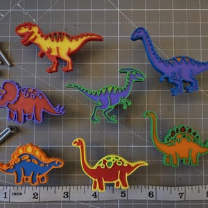 May include: A set of six colorful dinosaur-shaped drawer knobs in yellow, red, purple, green, orange, and blue, accompanied by six silver metal mounting screws and a measuring tape showing a scale in inches for size reference.