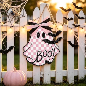 May include: A ghost-shaped wooden sign with a pink and white checkered pattern, featuring the black text "BOO!" and two black glittery bats attached. The sign is topped with a pink and black ribbon bow and hangs from a jute twine string.