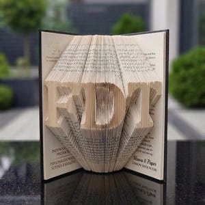 Könnte beinhalten: Eine gefaltete Buchskulptur mit den Buchstaben RDT, die durch präzise geknickte Buchseiten geformt wurden. Das offene Buch zeigt gedruckten Text auf den Innenseiten, einschließlich der Worte Słowa & Papir, was einen dreidimensionalen Effekt erzeugt.