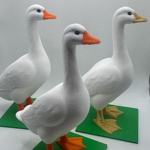 May include: Three white goose figurines standing on individual square green bases. Each goose has a textured white body, black eyes, and a distinct beak color. Two geese feature orange beaks and feet, while one goose has a gold-colored beak and yellow feet.