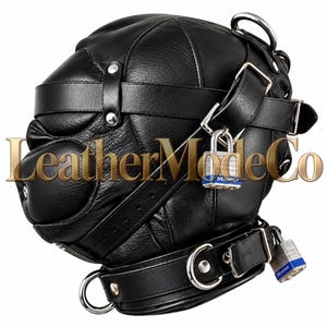 Könnte beinhalten: Eine schwarze Lederhaube mit mehreren verstellbaren Riemen, silberfarbenen Metallbeschlägen und zwei Master-Vorhängeschlössern mit blauen Akzenten. Der Text LeatherModeCo ist über die Mitte der schwarzen Lederkopfbedeckung gelegt.