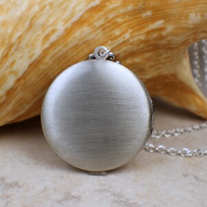 May include: A silver locket pendant with a brushed finish hangs from a silver chain. The locket is round and has a smooth surface.