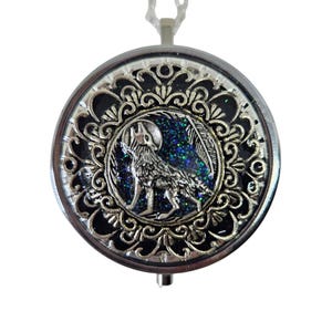 May include: Silver pendant necklace with a round, decorative pendant. The pendant showcases a detailed wolf howling at the moon, set against a dark blue glitter background. The pendant is suspended from a delicate silver chain.