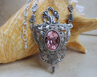 Puede incluir: Un collar con un colgante de plata con un diseño ornamentado y un gran cristal rosa. El colgante cuelga de una cadena de plata.