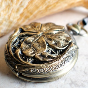 May include: A gold-toned pocket watch with a four-leaf clover design on the front. The watch has a detailed, ornate border.