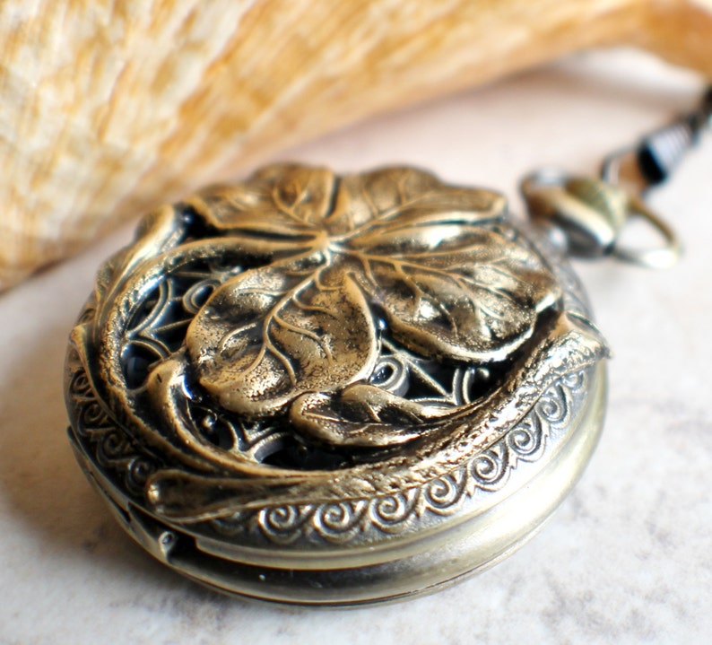 May include: A gold-toned pocket watch with a four-leaf clover design on the front. The watch has a detailed, ornate border.