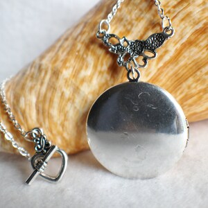 May include: A silver locket necklace with a delicate filigree design. The locket is round and has a hinged opening. The necklace is on a silver chain with a heart-shaped clasp.