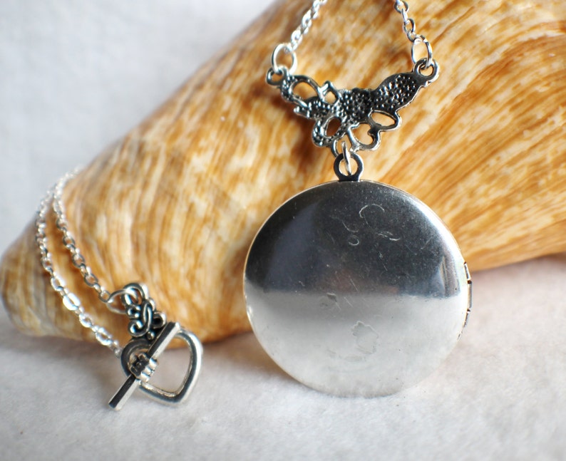 May include: A silver locket necklace with a delicate filigree design. The locket is round and has a hinged opening. The necklace is on a silver chain with a heart-shaped clasp.