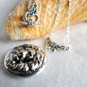 May include: A silver locket necklace with a lion head design. The necklace has a silver chain and a silver clasp with a heart-shaped design.