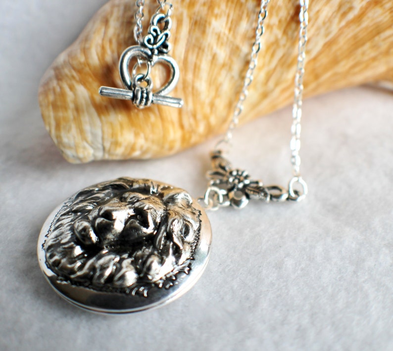 May include: A silver locket necklace with a lion head design. The necklace has a silver chain and a silver clasp with a heart-shaped design.