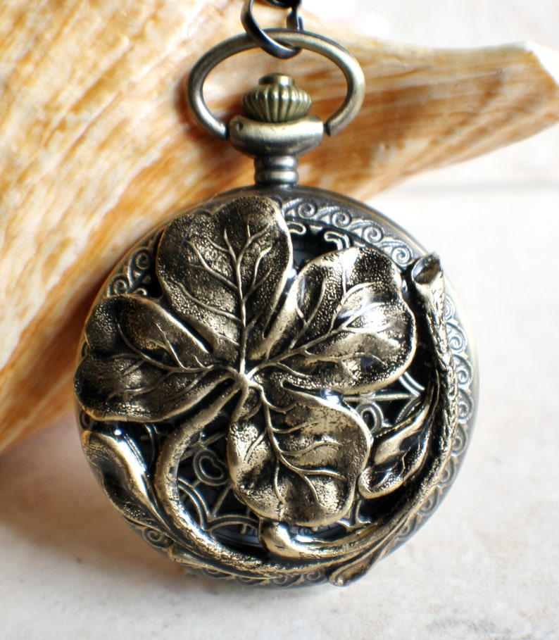 May include: A gold-toned pocket watch with a four-leaf clover design. The watch has a black chain and a decorative, ornate design around the edge.