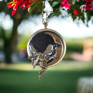 Può includere: Una collana con un ciondolo a forma di mezzaluna dorata su sfondo nero scintillante e una fata con ali iridescenti che vola davanti.