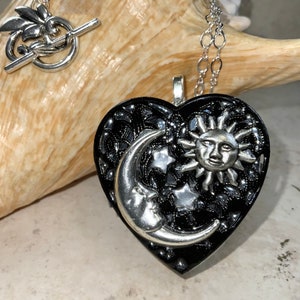 May include: A silver necklace with a black heart-shaped locket featuring a silver crescent moon and sun with a face. The locket is decorated with small silver stars.