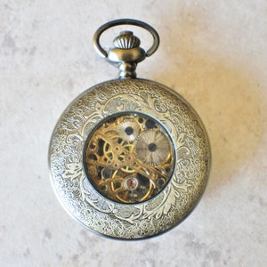 May include: A gold-toned pocket watch with an intricate floral design on the case. The watch face is open, revealing the inner workings of the mechanical movement.