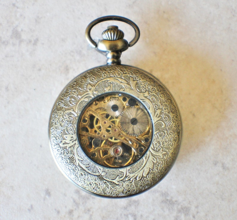 May include: A gold-toned pocket watch with an intricate floral design on the case. The watch face is open, revealing the inner workings of the mechanical movement.