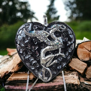 May include: A black heart-shaped pendant with a silver griffin design. The pendant has a detailed filigree pattern and a shiny finish.