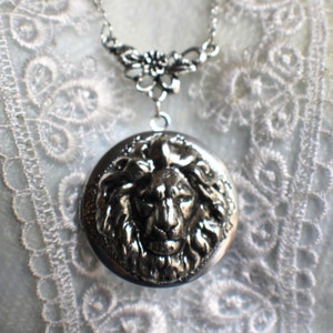 May include: Silver necklace with a locket featuring a detailed lion head design. The locket is attached to a delicate chain with a floral design.