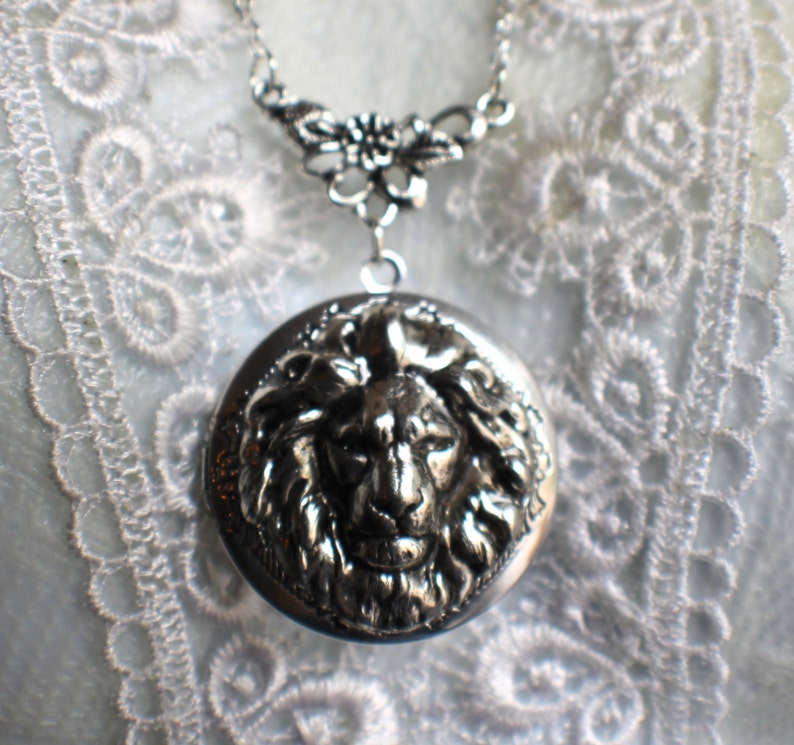 May include: Silver necklace with a locket featuring a detailed lion head design. The locket is attached to a delicate chain with a floral design.
