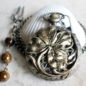 May include: A gold-toned pocket watch with a four-leaf clover design on the face. The watch is attached to a chain with brown beads.