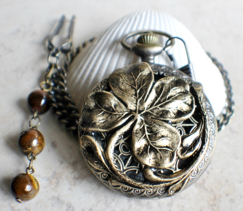 May include: A gold-toned pocket watch with a four-leaf clover design on the face. The watch is attached to a chain with brown beads.