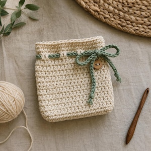 May include: A square, cream-colored crochet pouch featuring a textured stitch pattern. The pouch is cinched at the top with a sage green crocheted drawstring tied into a bow, accented by a single small, round, light brown wooden button.