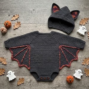 Puede incluir: Un pelele de manga larga tejido a ganchillo en color gris oscuro con paneles laterales en forma de alas de murciélago con detalles en naranja y morado, junto a un gorro a juego de ganchillo gris oscuro con orejas puntiagudas y tres botones.