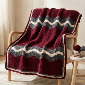 May include: A crocheted throw blanket featuring a chevron pattern in deep burgundy, charcoal gray, and cream. The blanket has a textured stitch and a border in matching colors. It is draped over a wooden chair with a small bowl of yarn nearby.