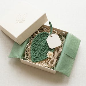 May include: A dark green crocheted leaf coaster nestled in a gift box with shredded paper filler, a blank white rectangular tag, a small dried flower, and a coil of twine. The box lid features a simple line drawing of two leaves.