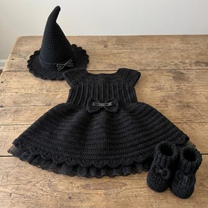 Puede incluir: Un conjunto negro de ganchillo que consta de un sombrero puntiagudo con borde festoneado y un pequeño lazo, un vestido de manga corta con cuerpo acanalado y falda acampanada con un lazo en la cintura, y un par de patucos a juego con lazos.