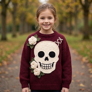 May include: A long-sleeved, deep burgundy knit sweater featuring a large, cream-colored crocheted skull appliqué on the front. The skull has black eye sockets and teeth. Three cream-colored crocheted roses with dark green leaves are attached.