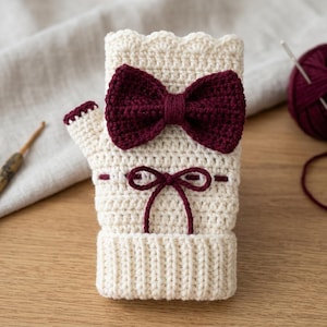May include: A cream-colored fingerless glove made of crochet yarn, featuring a large dark burgundy bow attached to the center. The glove has a scalloped top edge, a ribbed cuff at the bottom, and a small burgundy string tied in a bow below the main bow.