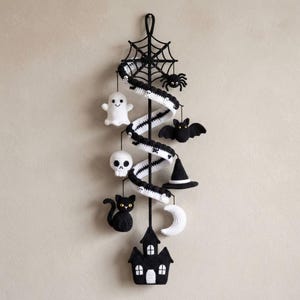 Könnte beinhalten: Ein vertikaler Wandbehang aus Häkelarbeit mit einem schwarzen Spinnennetz oben und einem spiralförmigen Band, das mit kleinen Totenköpfen verziert ist. An der Spirale hängen gehäkelte Figuren, darunter ein Geist, ein Totenkopf, eine schwarze Katze, eine Fledermaus, ein Hexenhut, ein Mond und ein Haus.