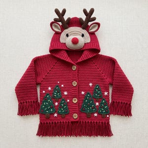 Op de afbeelding: Een rood gehaakt vest met capuchon, voorzien van een rendiergezicht op de capuchon met bruine geweien en een beige snuit. Het lijfje toont groene gehaakte kerstbomen met witte geborduurde sneeuwvlokken, houten knopen en lange franjes.