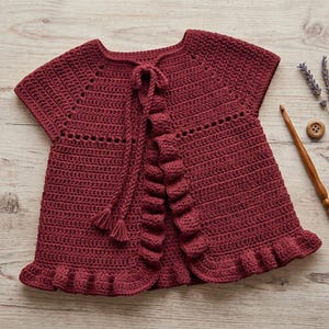 May include: A dark red crochet cardigan featuring short sleeves, a round neckline, and a front tie closure with tassels. The garment is detailed with a ruffled trim along the front opening and the bottom hem, showcasing a consistent stitch pattern.