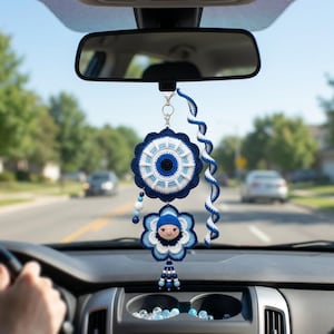 Può includere: Un ciondolo per auto all'uncinetto appeso allo specchietto retrovisore, caratterizzato da un motivo circolare a occhio di Allah in blu navy, azzurro e bianco, una piccola bambola con cappello blu, un cordino a spirale e una nappa di perline.