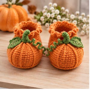 May include: A pair of orange crochet baby booties shaped like pumpkins. Each bootie features a ruffled top edge, a green crocheted stem, and a green leaf with embroidered details. The texture is consistent with yarn, showing a ribbed pumpkin design.