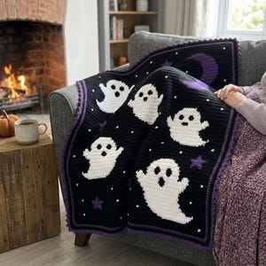Puede incluir: Una manta de ganchillo negra con figuras de fantasmas blancas, estrellas moradas y una luna creciente morada. La manta tiene un borde morado y pequeños detalles de punto de cruz blancos dispersos por el fondo oscuro.