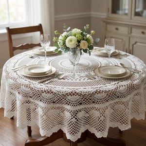 May include: A round white crochet tablecloth with an intricate openwork pattern featuring scalloped edges. The table is set with white ceramic plates, clear crystal wine glasses, and silver cutlery, centered with a bouquet of white flowers.